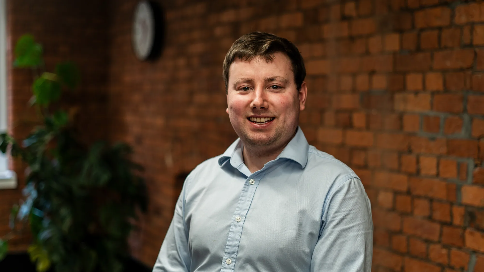 An individual in a light blue button‑up shirt sits indoors against a brick wall, with a clock on the wall and a large potted plant near a window in the background.
