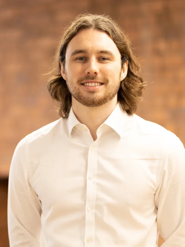 Darnell in an open collar white shirt, smiling to camera in front of a brick wall