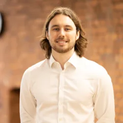 Darnell in an open collar white shirt, smiling to camera in front of a brick wall