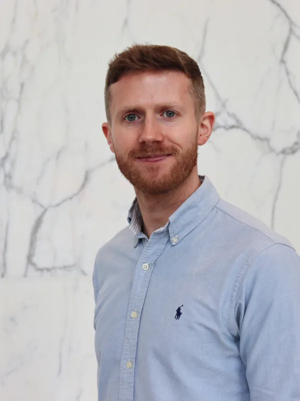 Portrait of a person standing against a white marble wall with grey veining, wearing a light blue button-down shirt with a small embroidered logo on the chest, facing the camera with a neutral expression in a softly lit indoor setting.