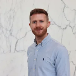 Portrait of a person standing against a white marble wall with grey veining, wearing a light blue button-down shirt with a small embroidered logo on the chest, facing the camera with a neutral expression in a softly lit indoor setting.