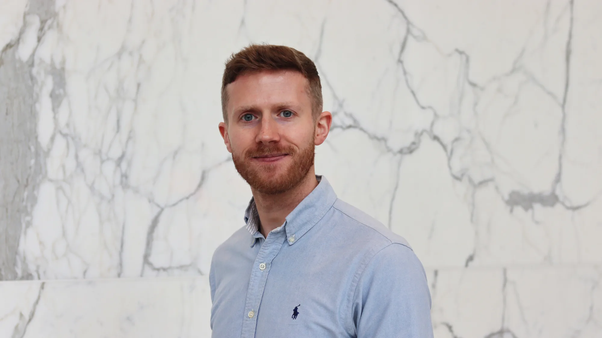 Portrait of a person standing against a white marble wall with grey veining, wearing a light blue button-down shirt with a small embroidered logo on the chest, facing the camera with a neutral expression in a softly lit indoor setting.
