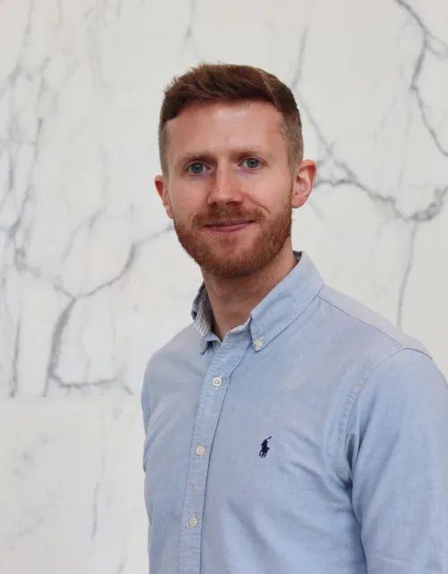 Portrait of a person standing against a white marble wall with grey veining, wearing a light blue button-down shirt with a small embroidered logo on the chest, facing the camera with a neutral expression in a softly lit indoor setting.