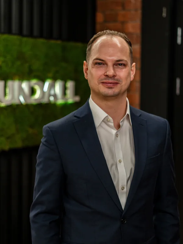 Joe wearing a dark suit jacket and white dress shirt stands indoors near a wall with a green moss panel displaying the text “CUNDALL,” with shelves and a doorway visible in the background.