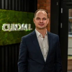 Joe wearing a dark suit jacket and white dress shirt stands indoors near a wall with a green moss panel displaying the text “CUNDALL,” with shelves and a doorway visible in the background.