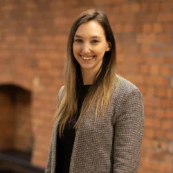 Kimberley stood smiling to camera in front of an exposed brick interior office wall