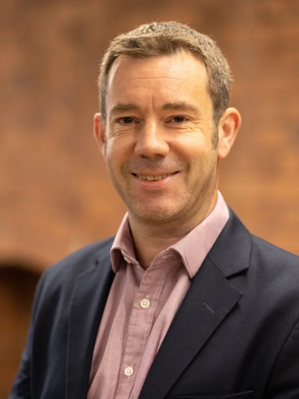 Head and sholders shot of Neil against brick wall background wearing pink shirt and blue blazer