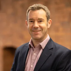 Head and sholders shot of Neil against brick wall background wearing pink shirt and blue blazer