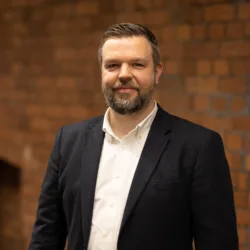 Peter Ridge in a white open collar shirt and suit jacket in front of a brick wall