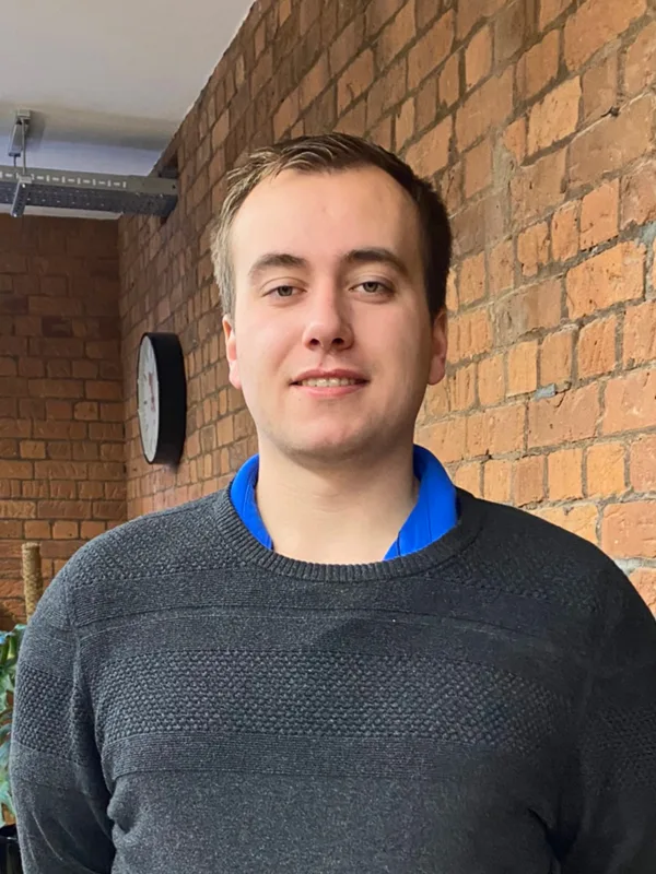 Sam Gardner wearing a grey jumper standing against a brick wall in the Manchester office