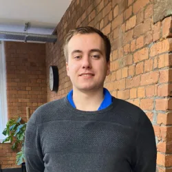 Sam Gardner wearing a grey jumper standing against a brick wall in the Manchester office
