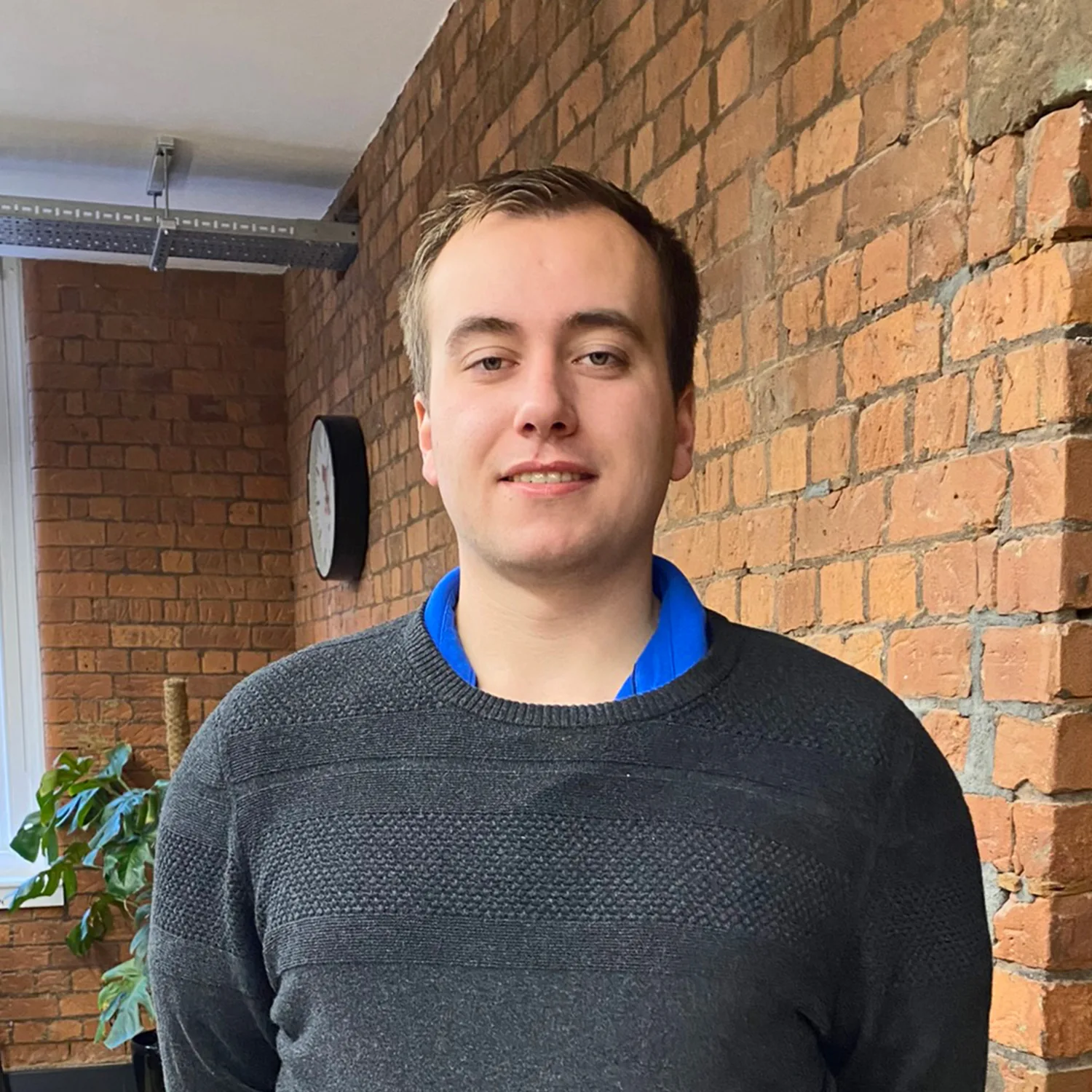 Sam Gardner wearing a grey jumper standing against a brick wall in the Manchester office