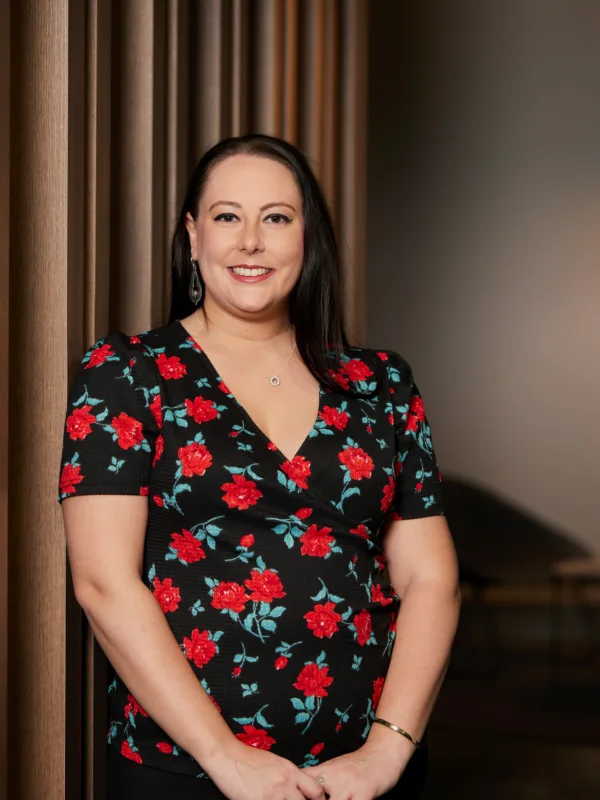 Alex in a dark top with red roses stood in front of a wooden panel backdrop