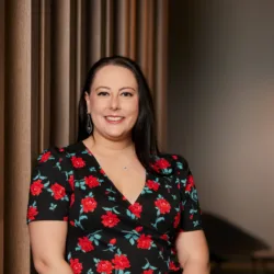 Alex in a dark top with red roses stood in front of a wooden panel backdrop