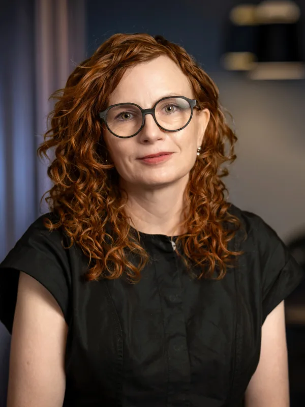 A person with shoulder-length curly red hair wearing a short-sleeved black top is seated indoors. The background shows dark vertical panels, two pendant lights, and blurred plants.