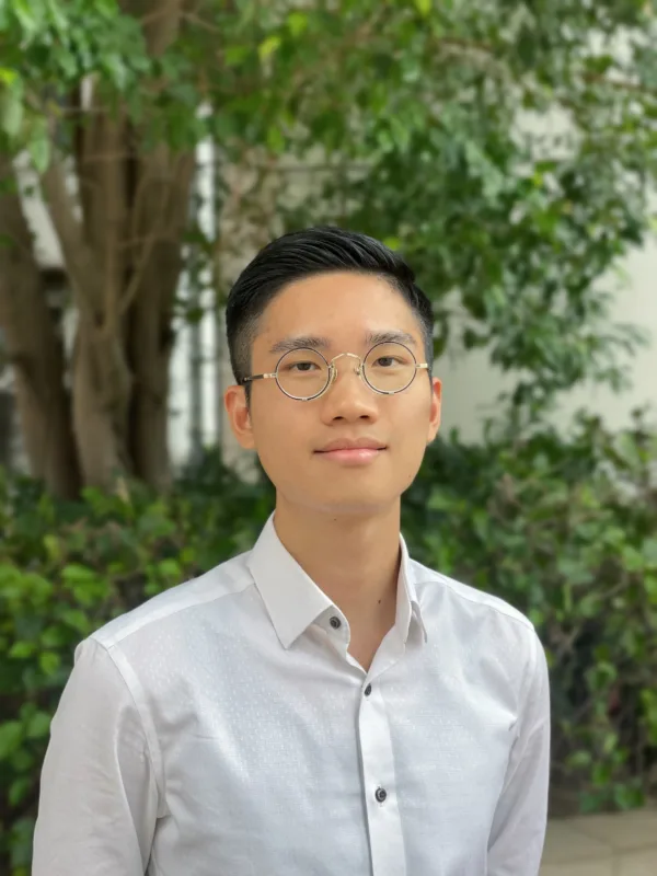 A person wearing round metal-framed glasses and a white button-up shirt is photographed from the chest up, standing outdoors in front of blurred green foliage and a tree trunk in soft natural light.