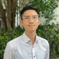 A person wearing round metal-framed glasses and a white button-up shirt is photographed from the chest up, standing outdoors in front of blurred green foliage and a tree trunk in soft natural light.