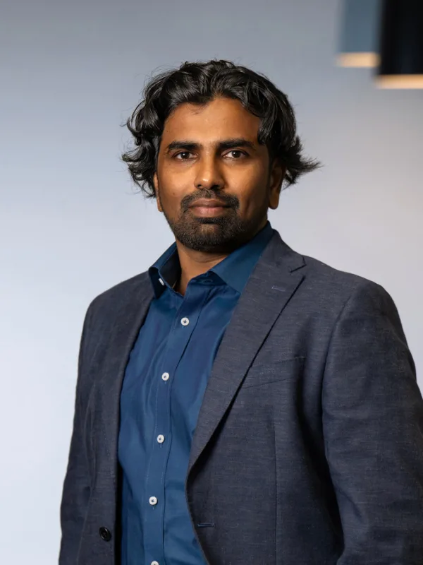Nanda wearing a dark blue shirt with a dark grey suit jacket standing with arms in pocket in an office setting