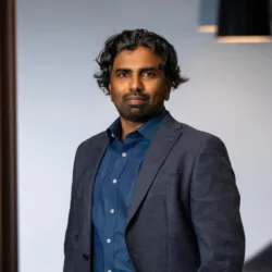 Nanda wearing a dark blue shirt with a dark grey suit jacket standing with arms in pocket in an office setting