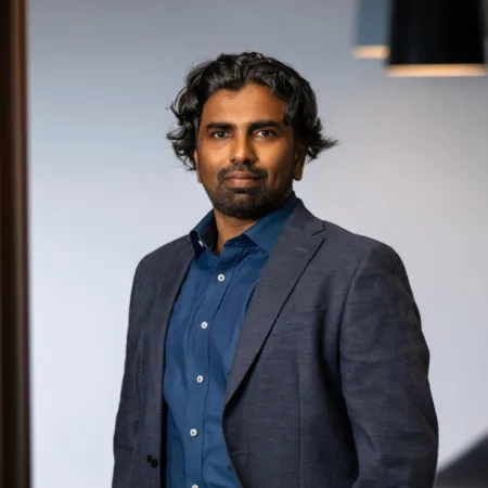 Nanda wearing a dark blue shirt with a dark grey suit jacket standing with arms in pocket in an office setting