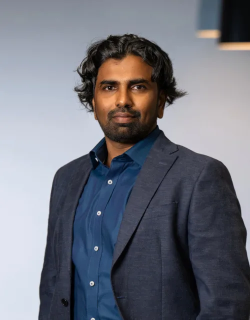 Nanda wearing a dark blue shirt with a dark grey suit jacket standing with arms in pocket in an office setting
