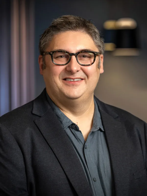 A man wearing a dark blazer over a buttoned shirt stands indoors against a softly lit background with dark curtains and hanging light fixtures