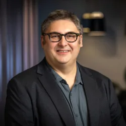 A man wearing a dark blazer over a buttoned shirt stands indoors against a softly lit background with dark curtains and hanging light fixtures
