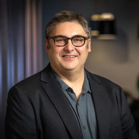 A man wearing a dark blazer over a buttoned shirt stands indoors against a softly lit background with dark curtains and hanging light fixtures