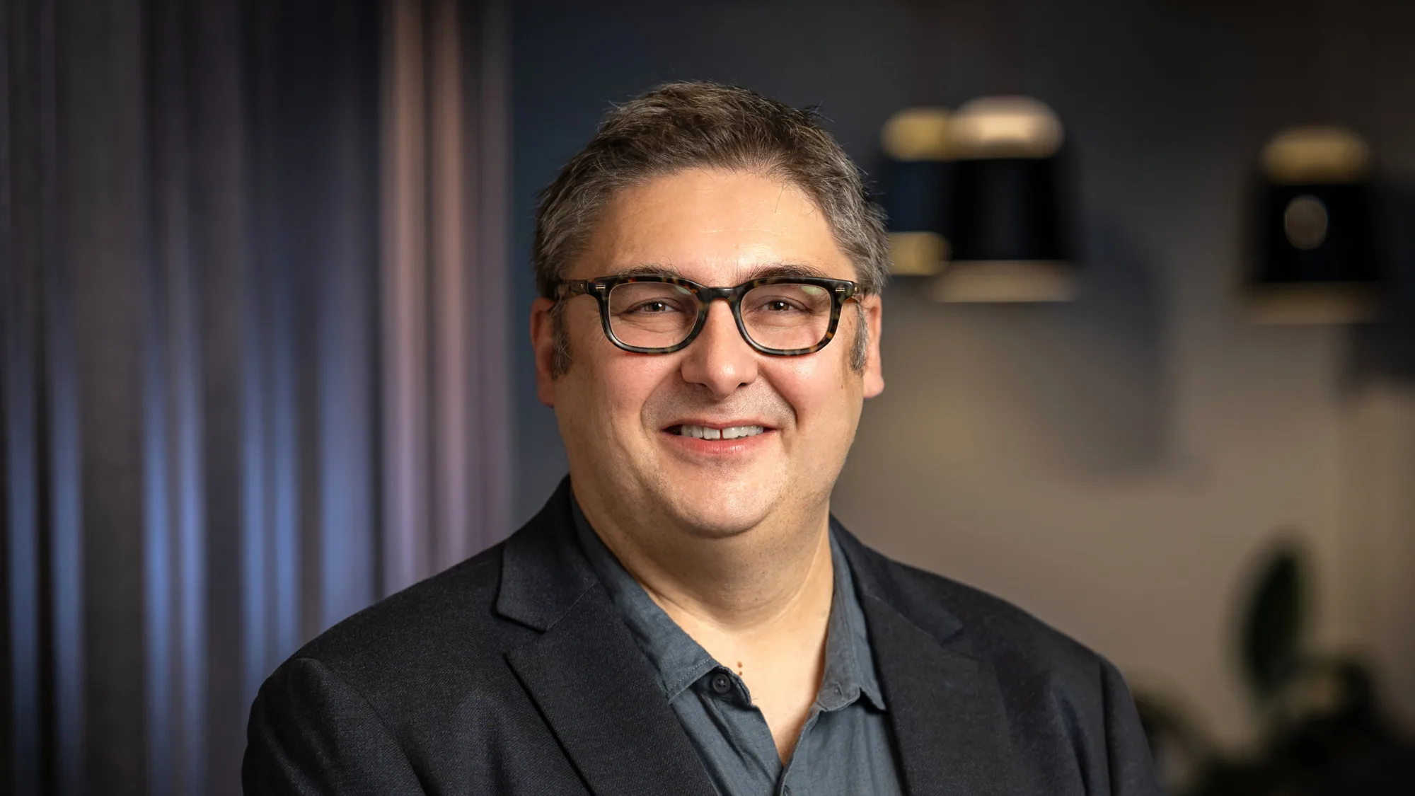 A man wearing a dark blazer over a buttoned shirt stands indoors against a softly lit background with dark curtains and hanging light fixtures