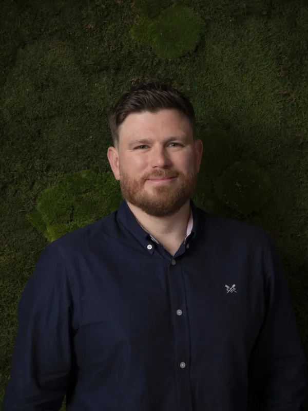 A person wearing a dark navy button-up shirt with a small embroidered logo on the left chest stands against a textured green moss wall background. The shirt is fully buttoned except for the top button.
