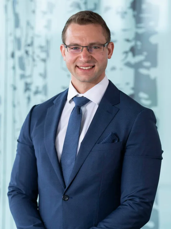 Athanasios wearing a dark suit, white shirt and blue tie, smiling to camera in front of a frosted glass screen.