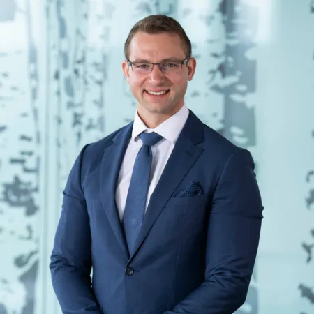 Athanasios wearing a dark suit, white shirt and blue tie, smiling to camera in front of a frosted glass screen.