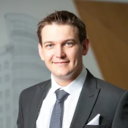 Barrie in a shirt tie and jacket smiling to camera in front of a vinyled office wall