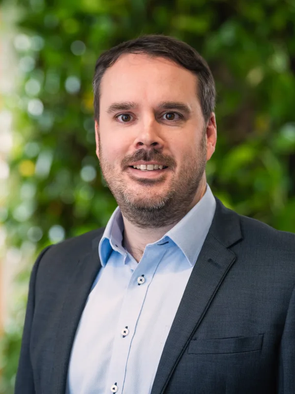 Colin looking to camera in a pale open collar shirt and suit jacket in front of a green plant wall