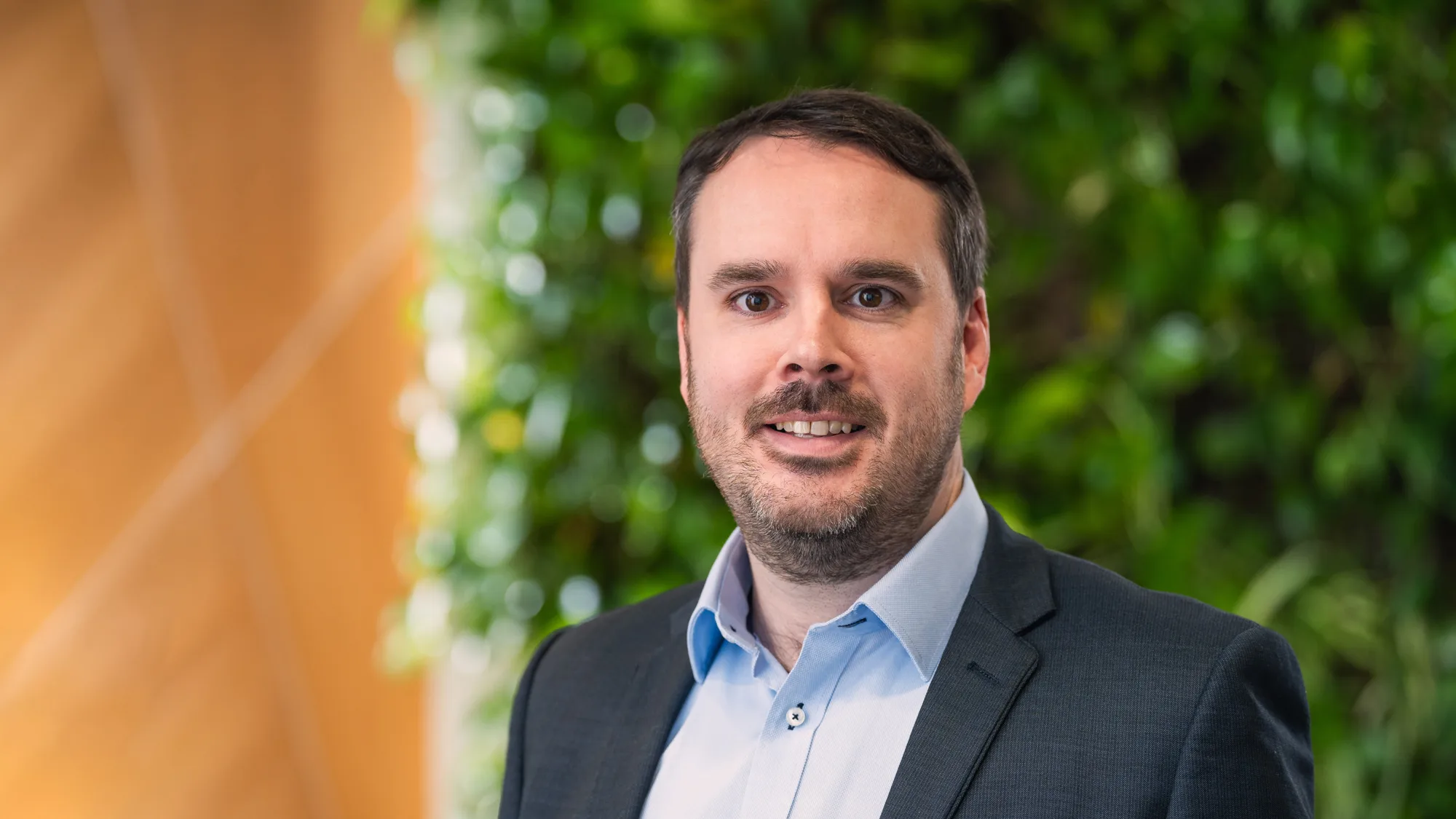 Colin looking to camera in a pale open collar shirt and suit jacket in front of a green plant wall