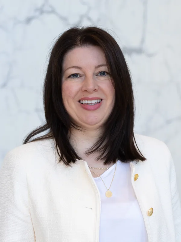 A person wearing a white textured jacket with gold buttons over a white top stands in front of a light grey marble wall. The jacket is open, and two layered necklaces with small pendants are visible.