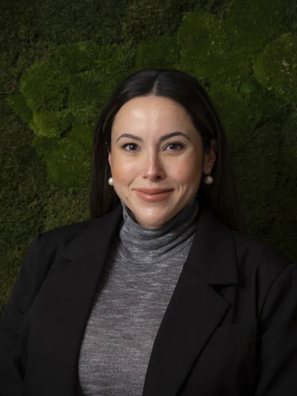A person wearing a dark blazer over a grey turtleneck top is positioned against a textured green wall with moss-like plants. The outfit includes pearl earrings visible on both sides.