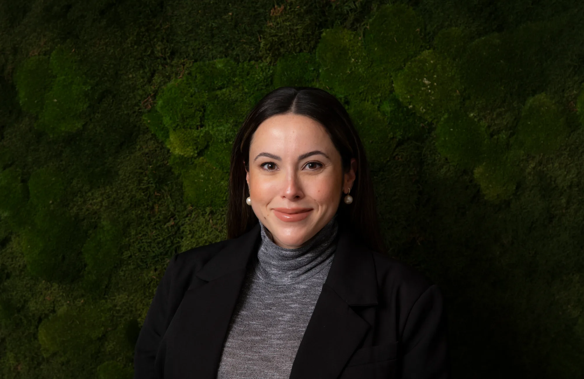 Portrait of a person wearing a dark blazer and grey turtleneck, standing against a textured green moss wall.