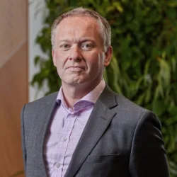Jim Allen in a grey suit and pink shirt on a wooden panelling and a living wall background
