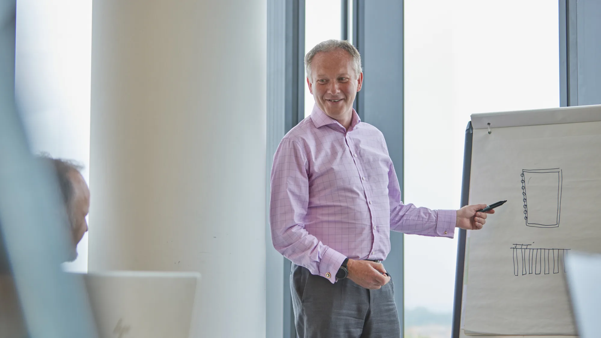 Jim Allen in a pink shirt and grey trousers pointing to flip chart in a meeting room with others