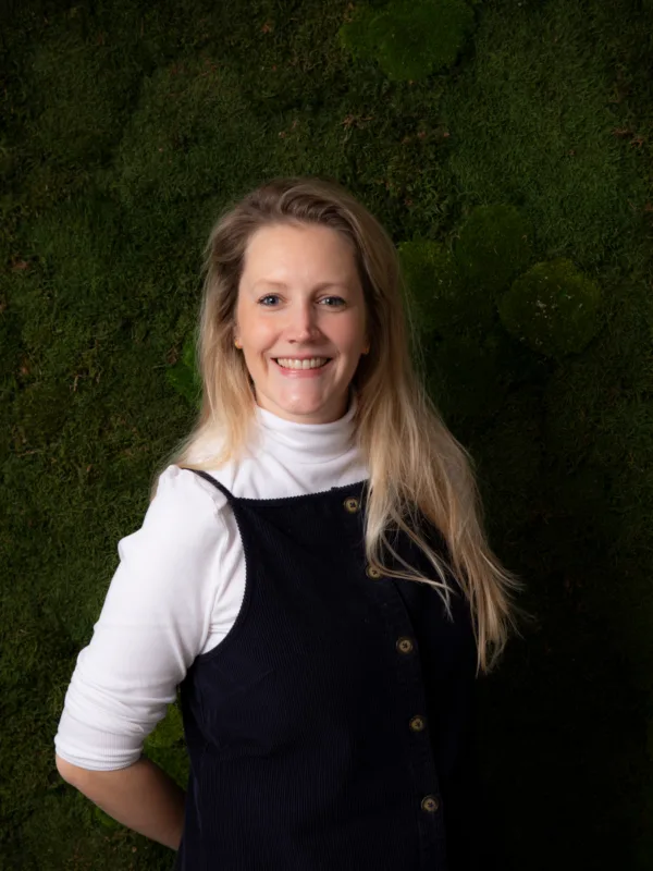 A person with long blonde hair stands against a textured green moss wall, wearing a white long‑sleeve top layered under a dark button‑front pinafore dress, with one arm positioned behind their back.