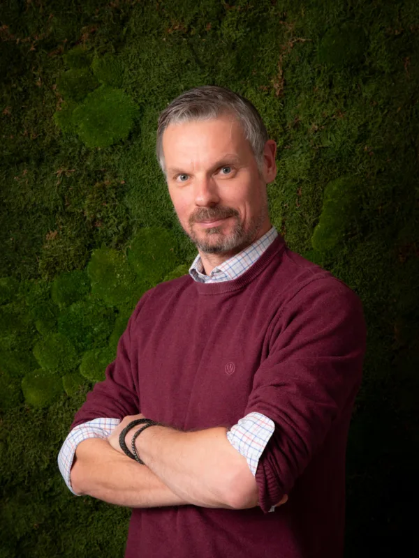A person wearing a burgundy sweater over a checked shirt stands with folded arms in front of a textured green moss wall.