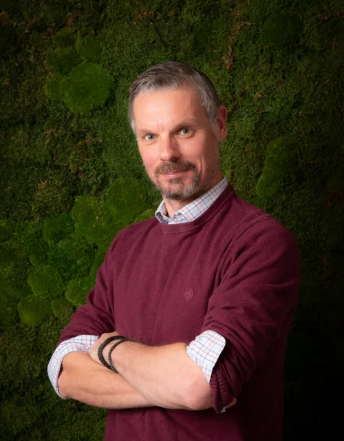A person wearing a burgundy sweater over a checked shirt stands with folded arms in front of a textured green moss wall.