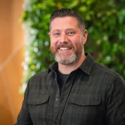 Head and shoulders shot of Oliver wearing a dark shirt against a green and brown background
