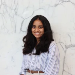 Portrait of a person with long dark hair wearing a blue and white striped shirt against a white marble wall.