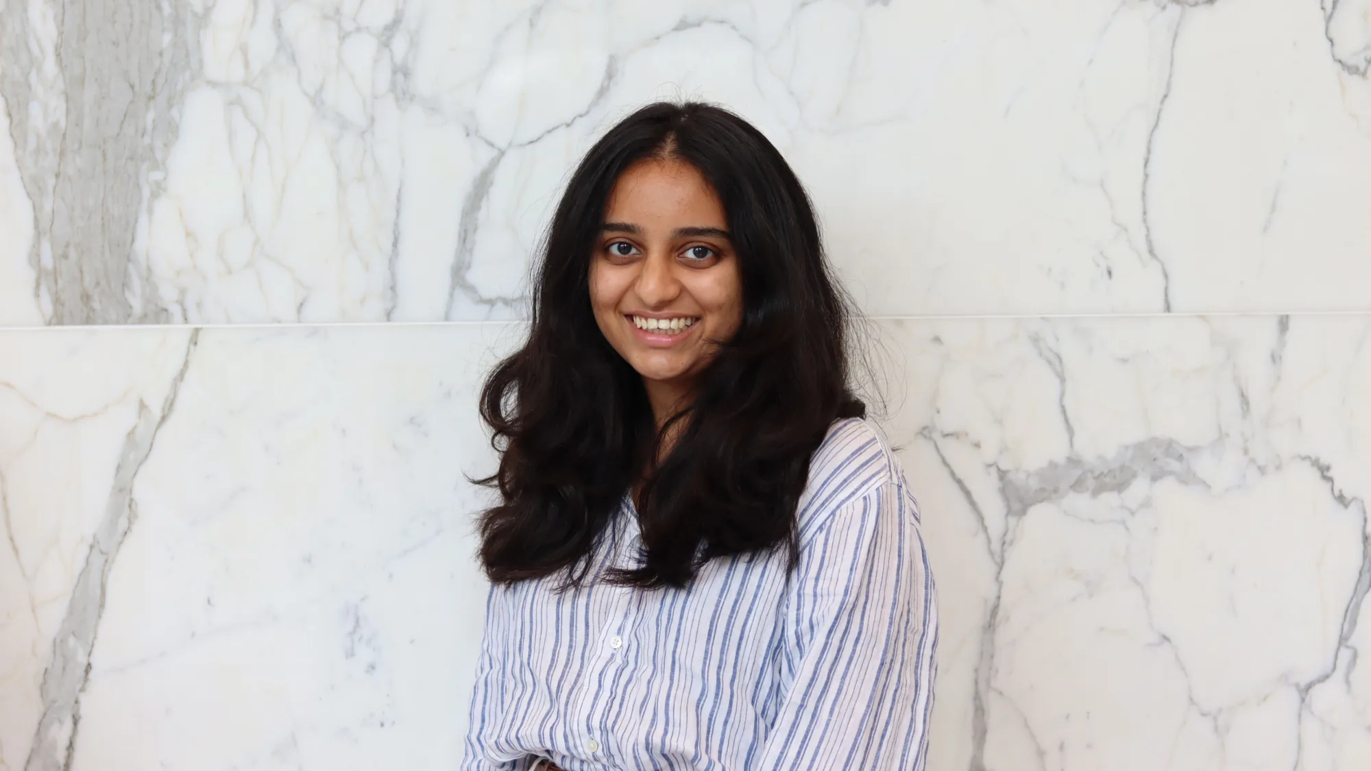 Portrait of a person with long dark hair wearing a blue and white striped shirt against a white marble wall.