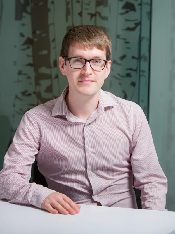 Stuart sat with his arm resting on a white desk in an open collar pale pink shirt and glasses in front of a frosted glass wall.