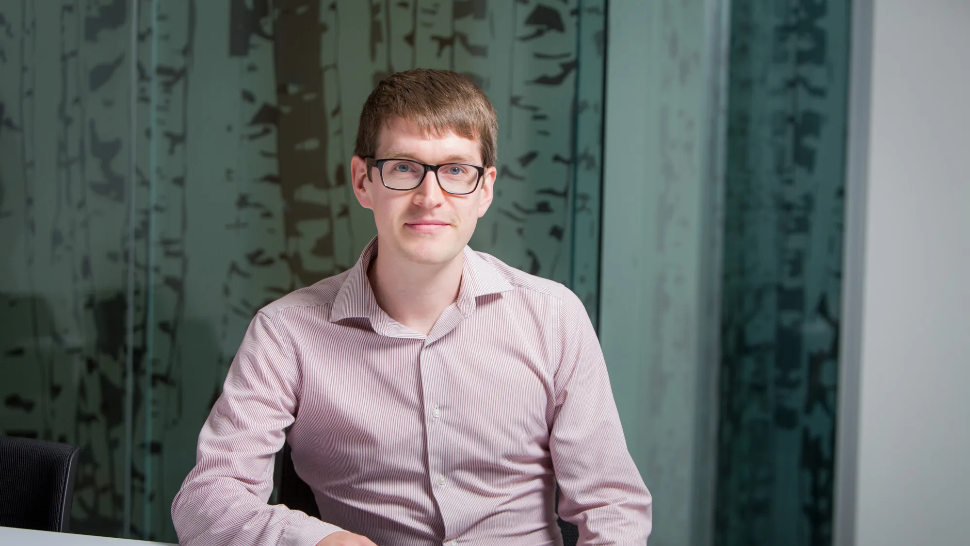 Stuart sat with his arm resting on a white desk in an open collar pale pink shirt and glasses in front of a frosted glass wall.