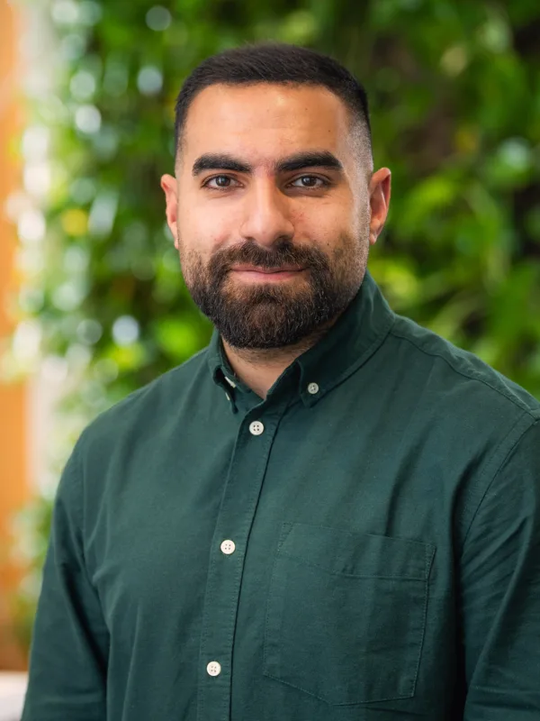 Person wearing a dark green button-up shirt stands in front of a background with green foliage and a wooden wall featuring diagonal lines.