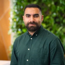 Person wearing a dark green button-up shirt stands in front of a background with green foliage and a wooden wall featuring diagonal lines.